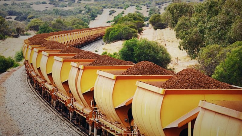 A long train of wagons loaded with iron ore curving through green countryside